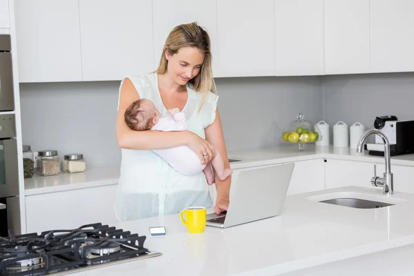 Мать использует ноутбук, неся ребенка на кухне — стоковое фото