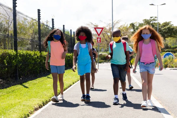 Multi ethnic group of school children wearing face masks, walking on the street. Education back to school health safety during Covid19 Coronavirus pandemic.