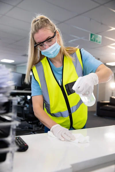 Caucasian woman sanitizing an office wearing hi vis vest, gloves, safety glasses and face mask, using disinfectant and wiping surfaces. Hygiene in workplace during Coronavirus Covid 19 pandemic.