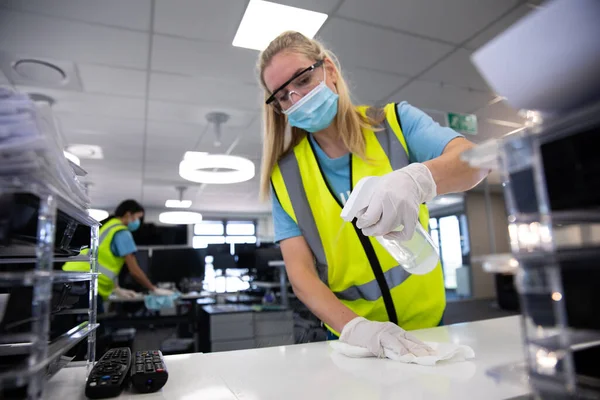 Caucasian woman and colleague wearing hi vis vests, gloves, safety glasses and face masks sanitizing an office using disinfectant. Hygiene in workplace during Coronavirus Covid 19 pandemic.