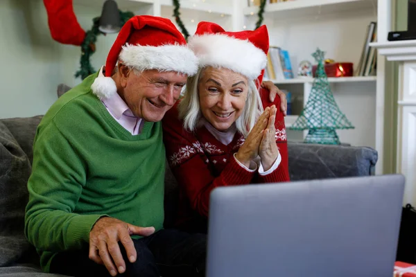 Senior couple in santa hats sitting on couch smiling while having a video call on laptop at home. social distancing during covid 19 coronavirus quarantine lockdown.