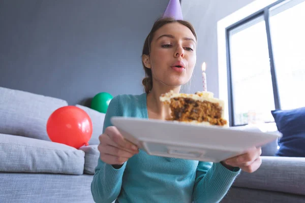 自宅で彼女の誕生日を祝う白人女性 パーティーの帽子をかぶってリビングルームで ケーキのスライスを保持し ろうそくを吹きます Covid 19コロナウイルス隔離ロックダウン中の社会的距離 — ストック写真