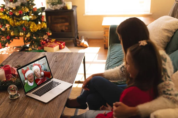 女人和女儿坐在沙发上 带着一个视频 家里的笔记本电脑上挂着头戴桑塔帽的老夫妇们笑着 圣诞佳节期间的社会疏离隔离观念 — 图库照片