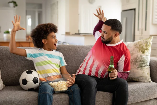 非洲裔美国人和他的儿子呆在家里 坐在客厅的沙发上 看电视上的运动节目 拿着爆米花和啤酒瓶 家庭一起在家共度美好时光 — 图库照片