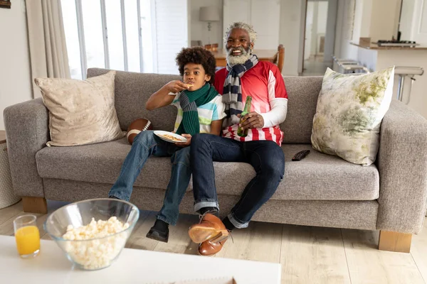 非洲裔美国人和他的孙子呆在家里 坐在客厅的沙发上 看电视上的运动节目 吃披萨 拿着啤酒瓶 家庭一起在家共度美好时光 — 图库照片