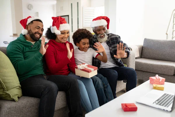 Multi-generation African American family at home sitting on a sofa in the living room, wearing Santa hats, waving at laptop during video chat. Family spending quality time at home together.