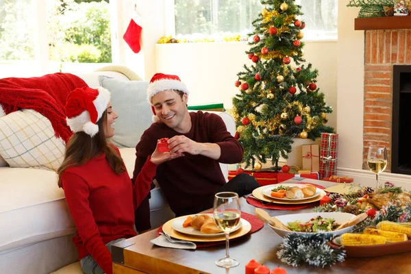 戴着桑塔帽的男人把礼品盒送给了家中客厅里的一个女人 圣诞节庆祝传统的概念 食环动物检疫期间的社会疏离 — 图库照片