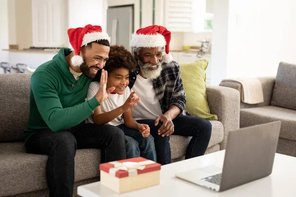 Multi-generation African American family at home sitting on a sofa in the living room, wearing Santa hats, waving at laptop during video chat. Family spending quality time at home together.