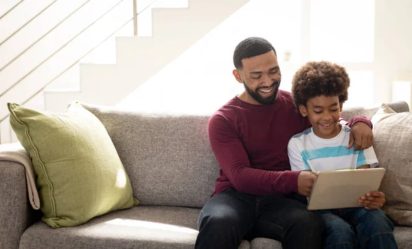 非洲裔美国人和他的儿子坐在客厅的沙发上 用平板电脑笑着 全家人都在家里慢吞吞地度过美好时光 — 图库照片