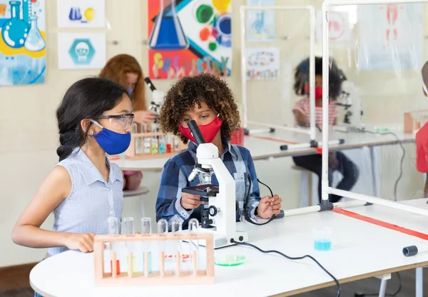 フェイスマスクをした女子高生2人が顕微鏡を見ている Covi19コロナウイルスパンデミックの際の学校の安全性への教育 — ストック写真