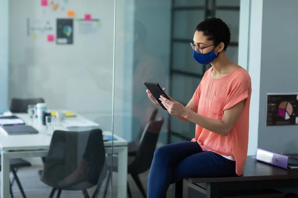 混血女子戴着面具坐在办公室里 使用数码平板电脑 大肠癌大流行期间工作场所的卫生情况D — 图库照片
