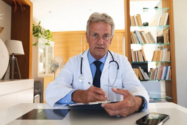 高级高加索男医生穿着实验室外套和听诊器坐在办公桌前 在视频通话中摆姿势拍照的画像 食管癌大流行期间的自我隔离 — 图库照片