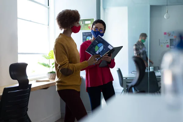 事務所では仮面をつけた2人の混合レース女性が話していた 文書を見て議論する同僚たちです コロナウイルスの職場での衛生は19のパンデミックを克服しました — ストック写真
