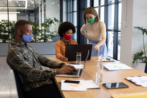 Diverse group of business people working in creative office. group of people wearing face masks and using laptops. social distancing protection hygiene in workplace during covid 19 pandemic.