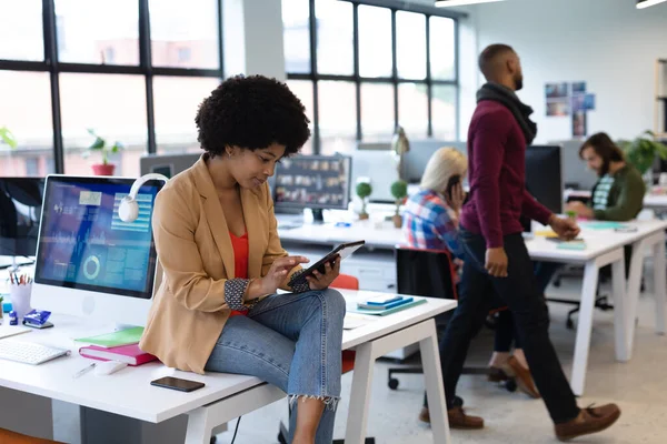 創造的なオフィスで働く混合レースビジネス女性 女性は机の上に座りタブレットPcを使っています ビジネス関係者や職場の同僚が — ストック写真