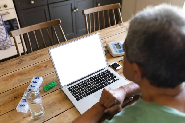 Senior african american woman with medication using laptop at kitchen table. retirement lifestyle in self isolation during coronavirus covid 19 pandemic.