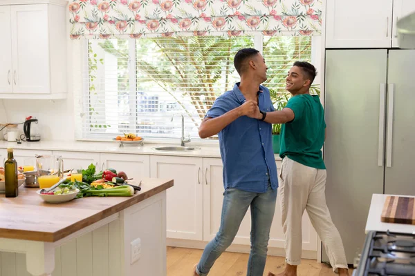 Diverse gay male couple spending time in kitchen dancing. staying at home in isolation during quarantine lockdown.