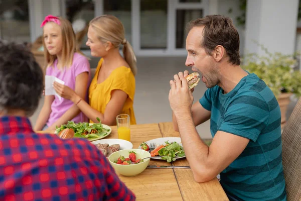 白人男性は庭で食事をする家族と一緒にテーブルに座ってハンバーガーを食べています 庭で食事をするテーブルに座る3世代の家族 — ストック写真