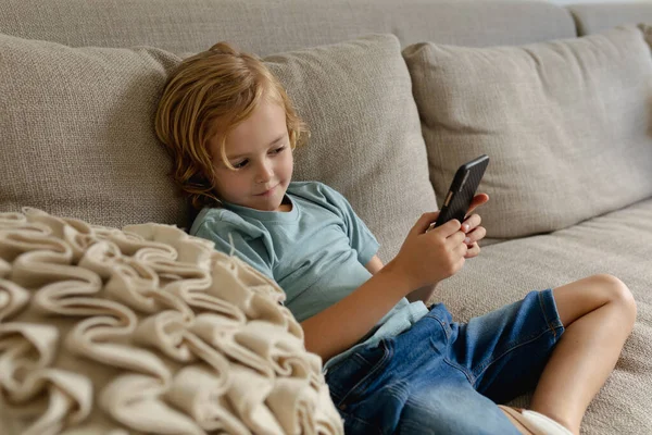 白种人男孩 一头长长的金发坐在沙发上 用智能手机 隔离检疫期间单独呆在家里 — 图库照片