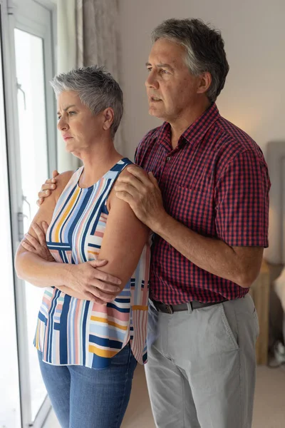 Concerned Senior Caucasian Couple Standing Looking Out Window Embracing Staying — Stock Photo, Image