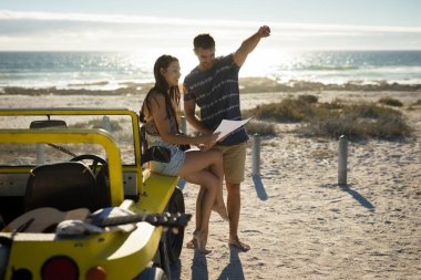 Happy caucasian couple sitting on beach buggy by the sea reading road map pointing. beach break on summer holiday road trip. clipart