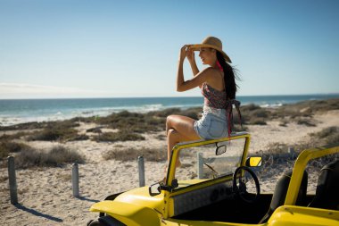 Happy caucasian woman sitting on beach buggy by the sea wearing straw hat looking toward sea. beach break on summer holiday road trip. clipart