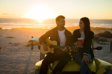 Happy caucasian couple sitting on beach buggy by the sea playing guitar and drinking during sunset. beach break on summer holiday road trip. clipart