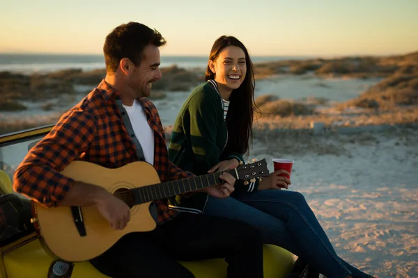 一对快乐的高加索夫妇坐在海边的童车上 一边弹吉他 一边在夕阳西下喝酒 暑假期间的海滩休息 — 图库照片