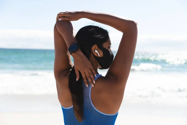混合赛女子戴着面罩和无线耳机在海滩上伸展胳膊进行运动 食管癌大流行期间海上健康的户外闲暇时间 — 图库照片