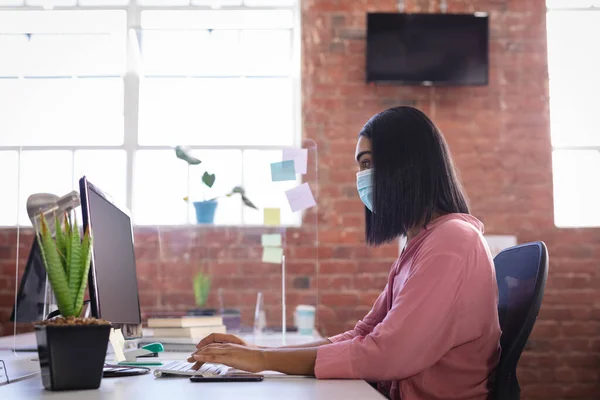 Yüz Maskesi Takmış Melez Bir Kadını Ofiste Bilgisayarın Önünde Oturuyor — Stok fotoğraf