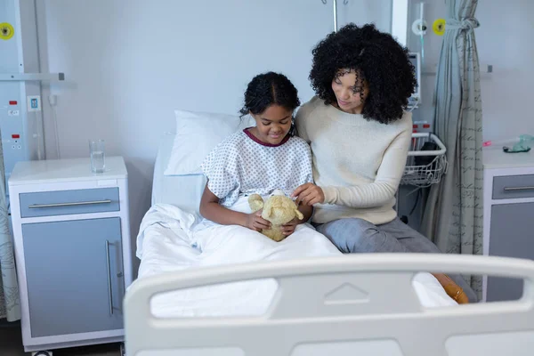 病院のベッドでテディベアを抱えている病気の娘を慰める混合レースの母親 医療サービス — ストック写真