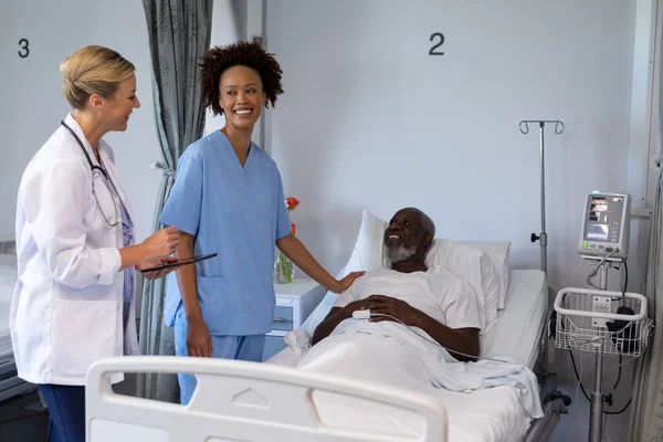 病院の部屋で2人の多様な女性医師とアフリカ系アメリカ人男性患者が笑顔で話しています 医療サービス — ストック写真