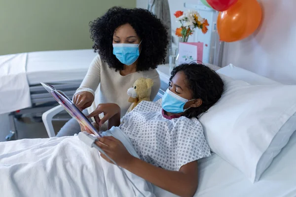 混合种族的母亲和生病的女儿在医院的面具 阅读书籍与泰迪熊 眼镜蛇大流行期间的医疗 保健和保健服务D — 图库照片