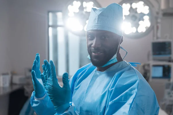 Glimlachende Afro Amerikaanse Mannelijke Chirurg Met Gezichtsmasker Beschermende Kleding Operatiekamer — Stockfoto