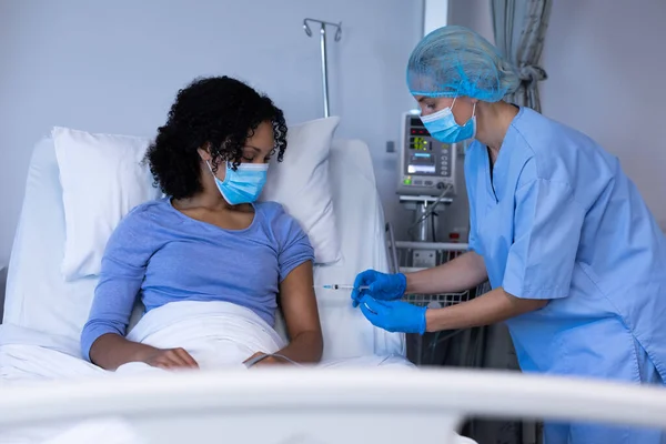 アフリカ系アメリカ人女性患者を予防接種するフェイスマスクを着用した病院の白人女性医師 コロナウイルス感染症の治療に従事する医療従事者は — ストック写真