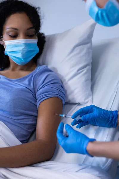 アフリカ系アメリカ人女性患者を予防接種するフェイスマスクを着用した病院の白人女性医師 コロナウイルス感染症の治療に従事する医療従事者は — ストック写真