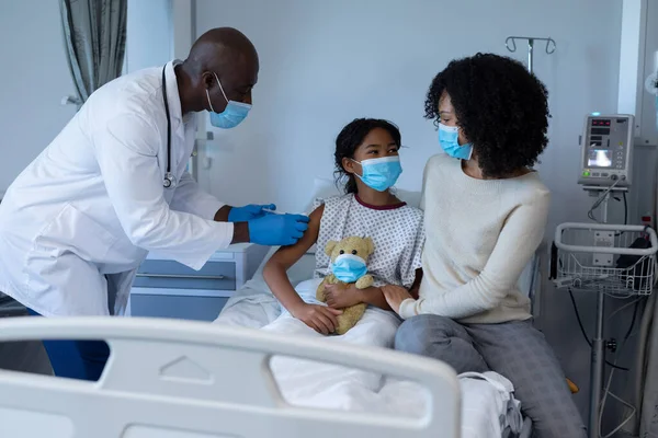 男性医師は すべての顔のマスクで 母親と病院のベッドで病気の女の子に快活な予防接種を与える コロナウイルスにおける医療 医療サービス Covid 19パンデミック — ストック写真