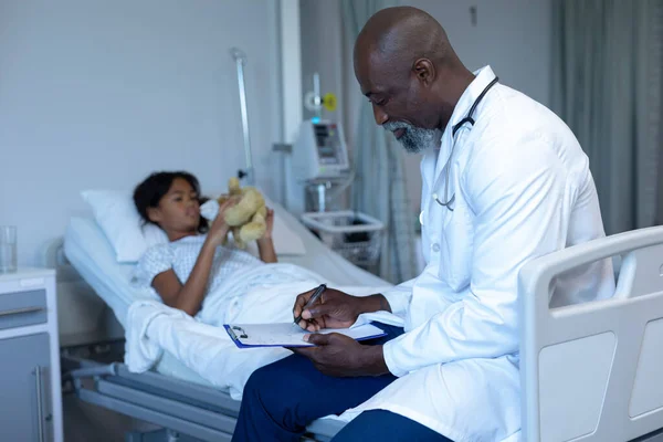 Médico Afro Americano Escrevendo Notas Sentadas Leito Hospitalar Paciente Mestiço — Fotografia de Stock