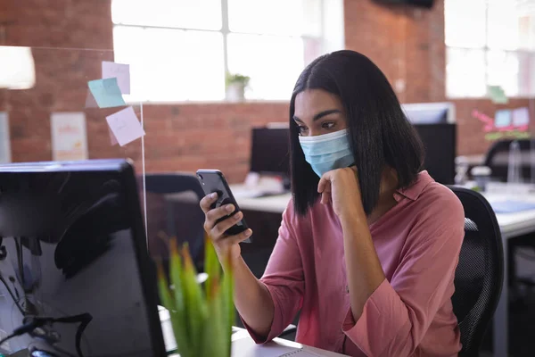 Ofiste Oturan Melez Kadını Akıllı Telefon Kullanarak Yüz Maskesi Takıyor — Stok fotoğraf