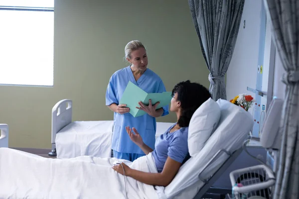 混合人種の患者と話をファイルを保持白人女性医師は 病院のベッドに座っている 医療サービス — ストック写真