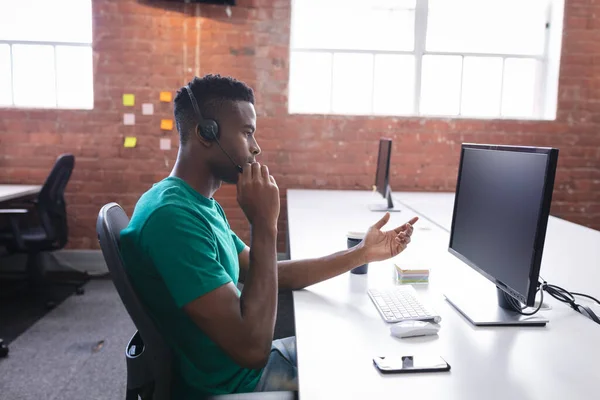 ヘッドフォンを使用してコンピュータの前に座っているビデオ通話を持つアフリカ系アメリカ人ビジネスマン 独立した創造的なデザインビジネス — ストック写真