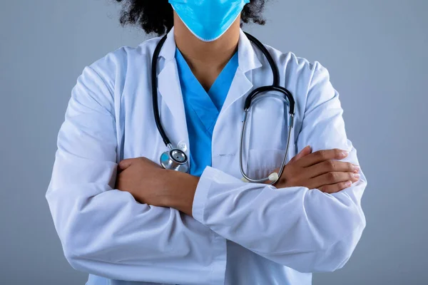 灰色の背景に腕を組んで顔マスクをした女医の真ん中部分が交差 医療と医療のプロフェッショナリズムの概念 — ストック写真