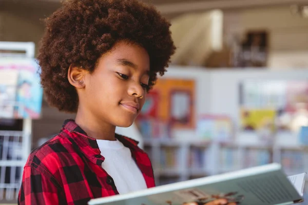 アフリカ系アメリカ人の少年が学校の図書館に立って本を読んで笑っている 子供の頃と小学校での教育は — ストック写真