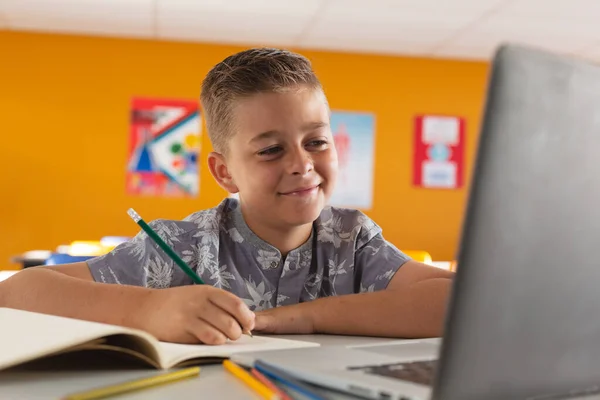 白人男孩坐在教室写字台前 看着笔记本电脑 面带微笑 小学的儿童 技术和教育 — 图库照片
