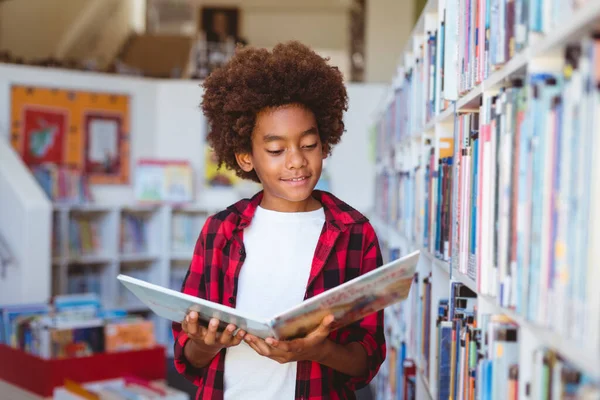 アフリカ系アメリカ人の少年が学校の図書館に立って本を読んで笑っている 子供の頃と小学校での教育は — ストック写真