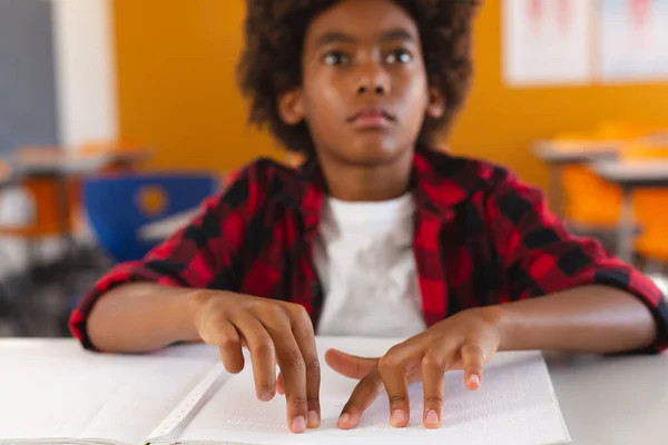 アフリカ系アメリカ人の視覚障害者の少年が教室の机に座り 指で点字本を読んでいます 子供の頃と小学校での教育は — ストック写真