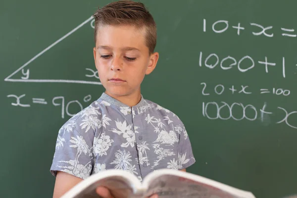 在数学课期间 白人男孩站在教室的黑板前拿着课本 儿童与小学教育 — 图库照片