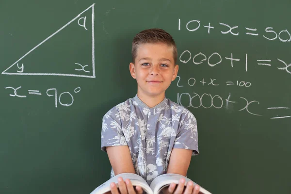 数学教室で読書をしながら黒板に立っている幸せな白人少年の姿 子供の頃と小学校での教育は — ストック写真
