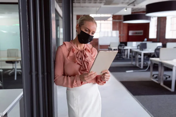タブレットを使用してオフィスに顔マスクを身に着けている白人実業家 コロナウイルスの流行期に近代的なオフィスで仕事をしていました — ストック写真