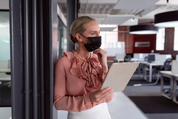 タブレットを使って事務所に顔マスクをしている思慮深い白人実業家は 目を背けている コロナウイルスの流行期に近代的なオフィスで仕事をしていました — ストック写真